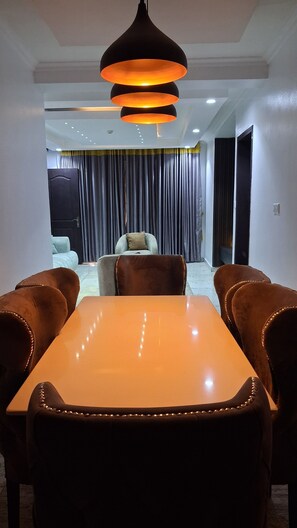 Un coin repas avec une table et des chaises, un luminaire suspendu et une vue sur un salon comprenant un canapé et des rideaux.
