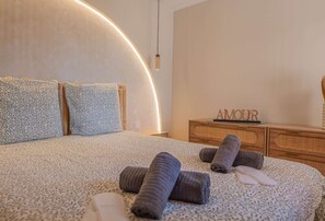 Ein modernes Schlafzimmer mit einer gebogenen Kopfteil, einer Holzkommode und gerollten Handtüchern auf dem Bett.