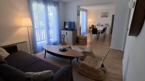 Uma sala de estar moderna com um sofá, uma mesa de centro e uma televisão. Há uma área de jantar com cadeiras e uma mesa ao fundo.