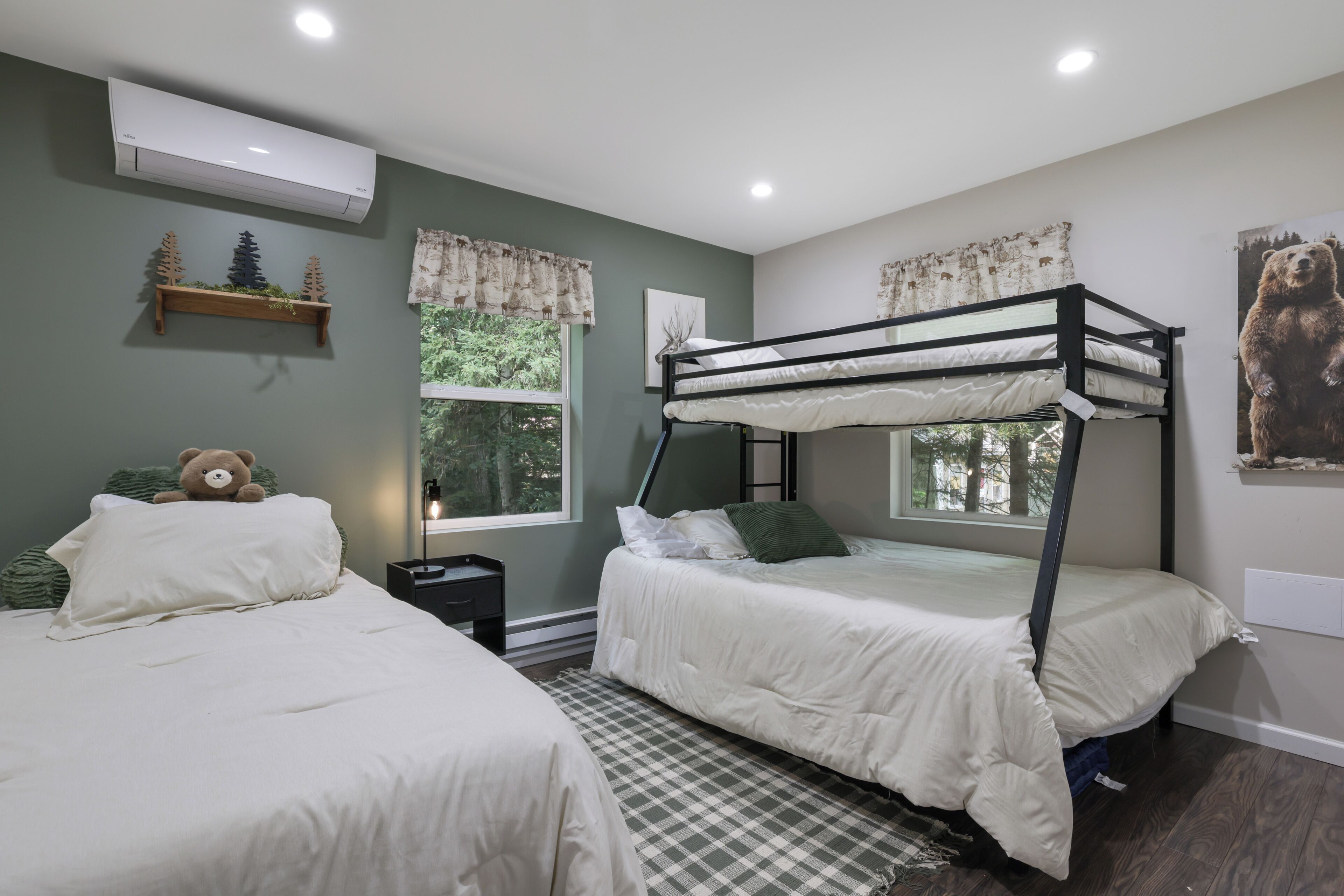 A bedroom with two bunk beds, a window with a view of trees, a wall-mounted air conditioner, and a bedside lamp.