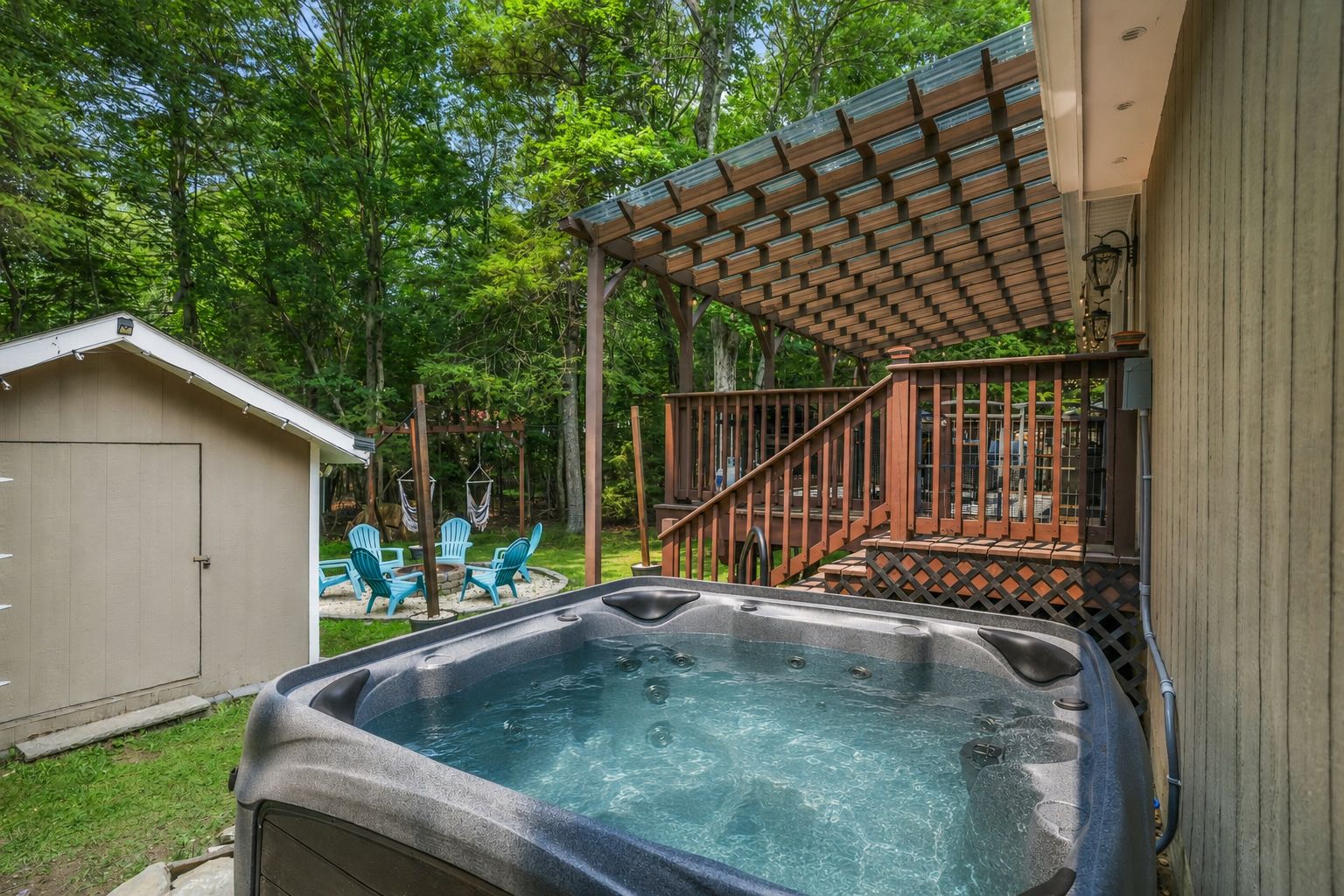 A hot tub with a wooden deck and a pergola, surrounded by trees and a small shed.