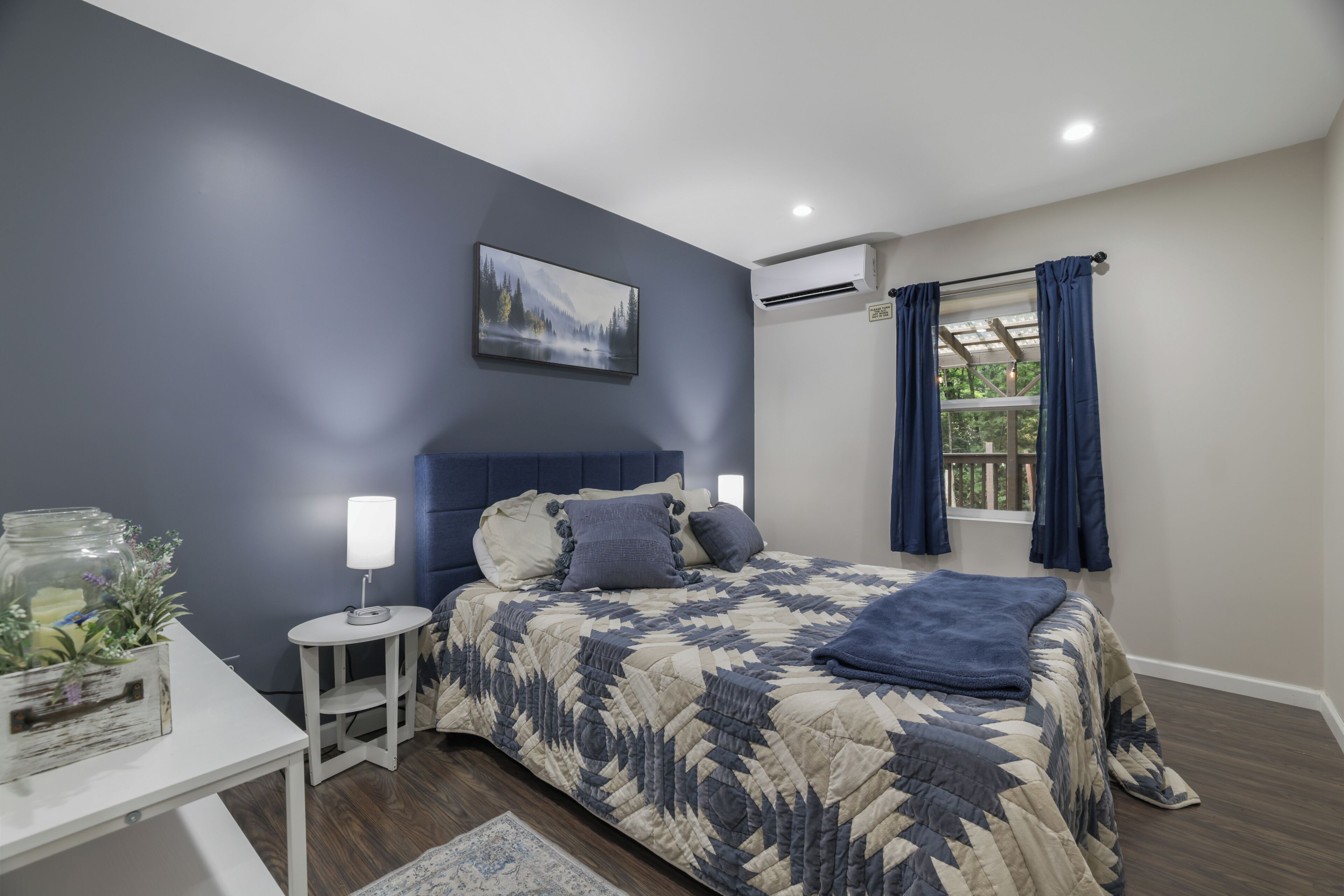A bedroom with a bed, bedside tables, a nightstand, a window with curtains, and an air conditioning unit.