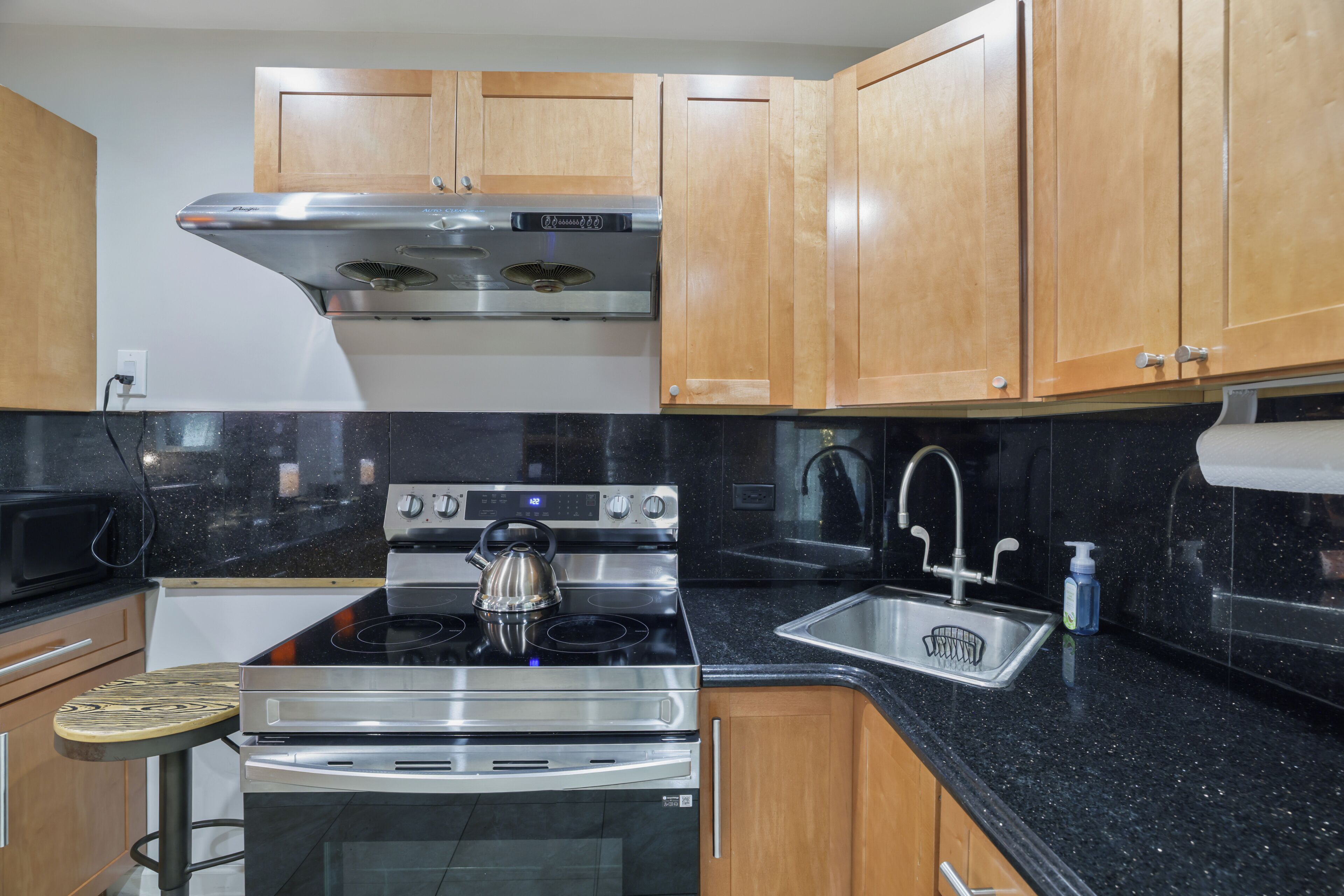 A kitchen with wooden cabinets, a stainless steel range hood, a kettle on the stove, a microwave, and a sink with a soap dispenser.