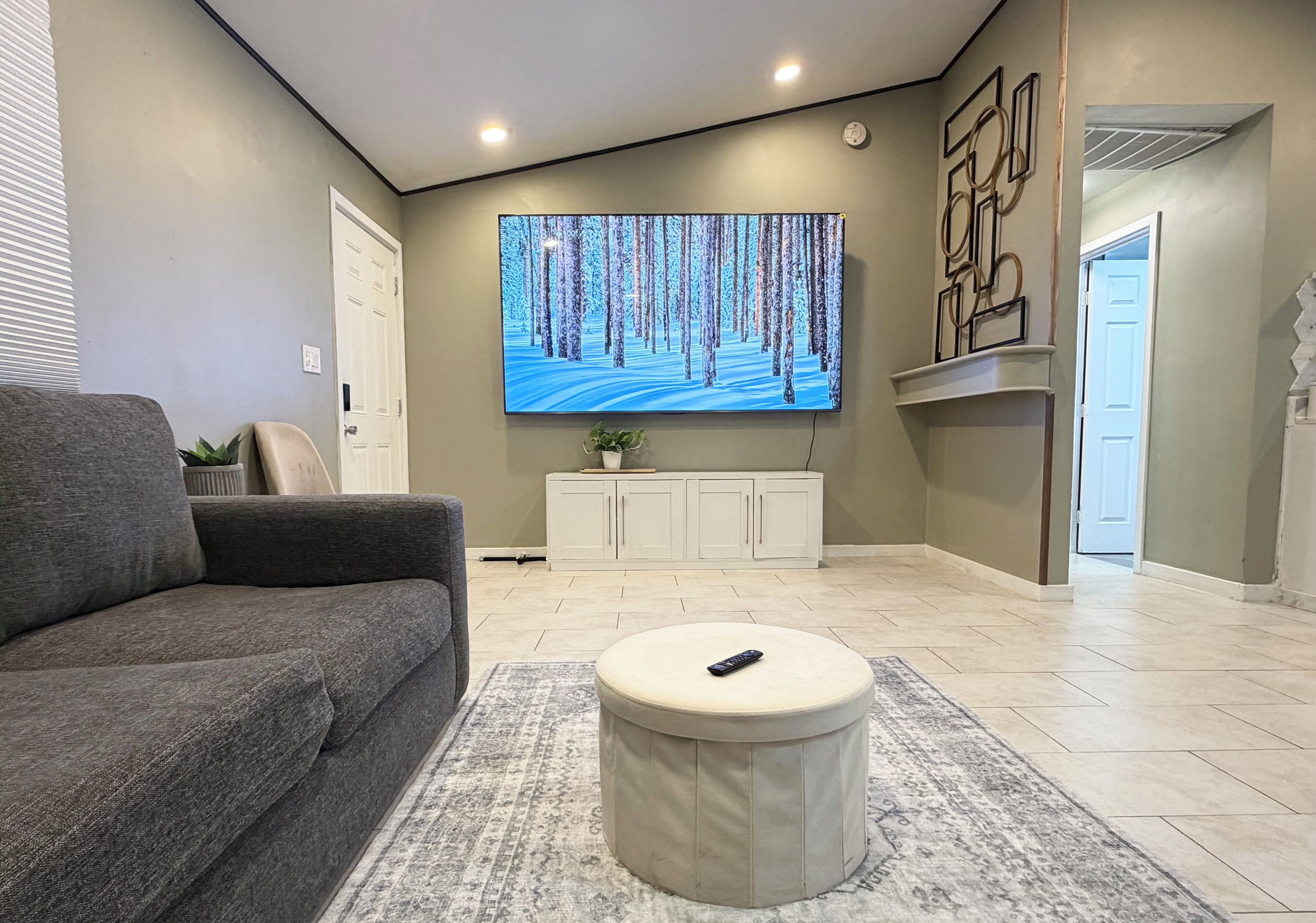 A living room with a grey sofa, a round ottoman, a flat-screen TV on the wall, a cabinet, and a door.