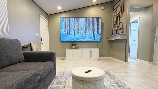 A living room with a grey sofa, a round ottoman, a flat-screen TV on the wall, a cabinet, and a door. -  Modern 2BR • 10 Min to Sky Harbor & Downtown (Phoenix)