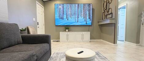 A living room with a grey sofa, a round ottoman, a flat-screen TV on the wall, a cabinet, and a door.