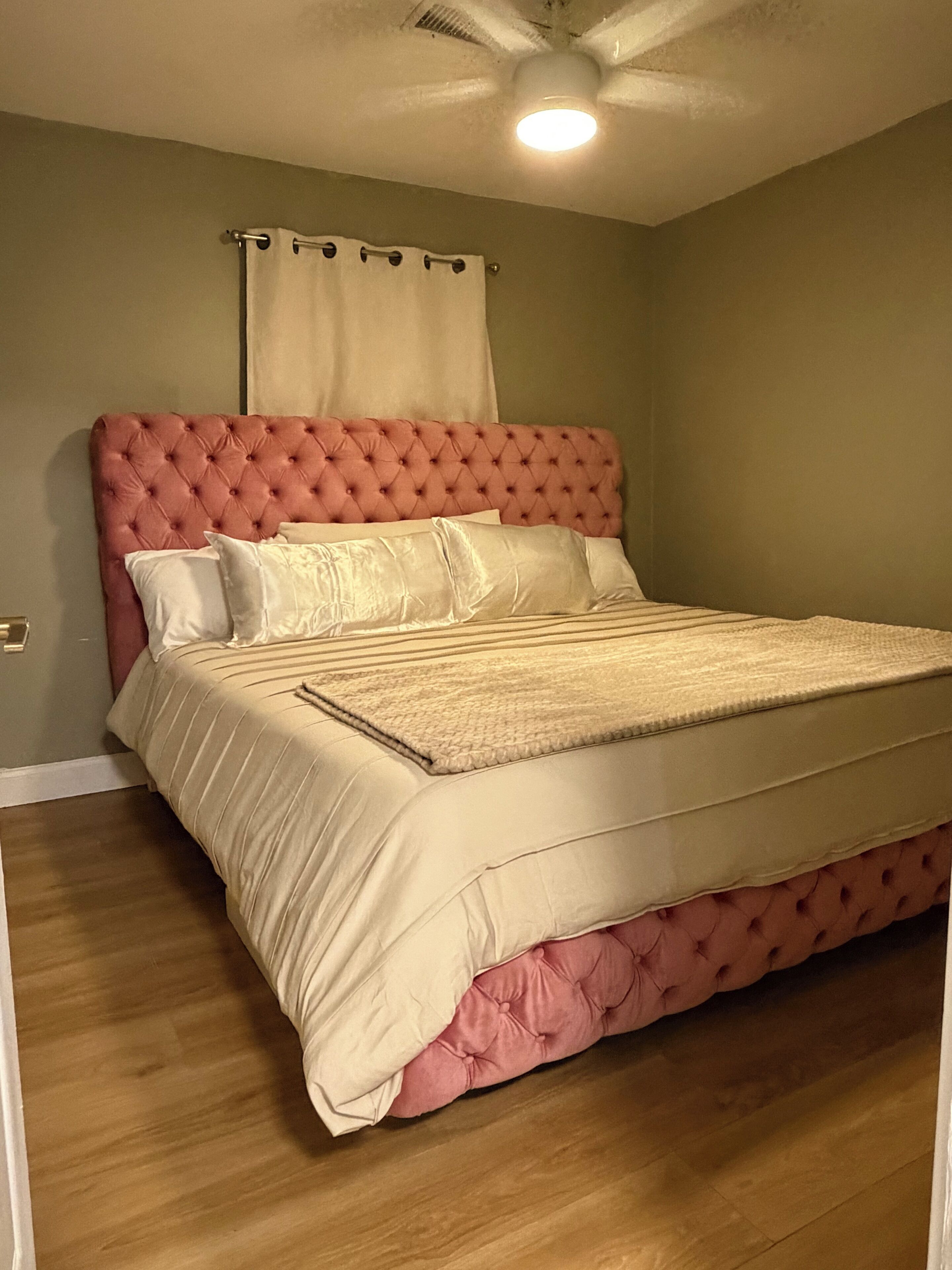 Une chambre à coucher avec une tête de lit capitonnée, un grand lit recouvert d’une couverture beige et un ventilateur de plafond.