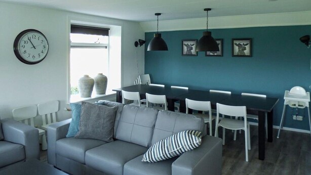 A modern living room with a grey sofa, a dining table with chairs, a clock, and a window with vases. - 5 bedroom nice home in Schalkhaar (Schalkhaar)