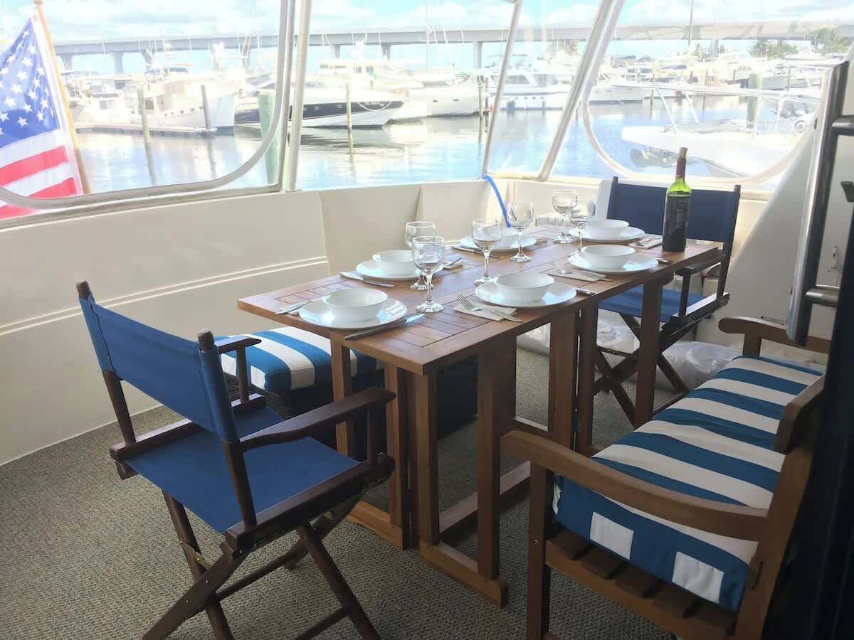 A dining area with a table set for four, chairs with blue cushions, and a view of boats through large windows.