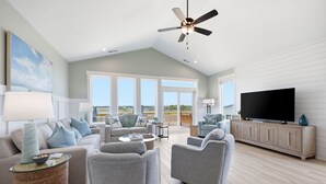 Ein geräumiges Wohnzimmer mit Deckenventilator, großen Fenstern, einem Flachbildfernseher und Holzboden.