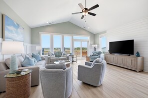 Ein geräumiges Wohnzimmer mit Deckenventilator, großen Fenstern, einem Flachbildfernseher und Holzboden.