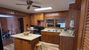 Una cocina con armarios de madera, una isla central con un taburete, un ventilador de techo y una pared de ladrillo.