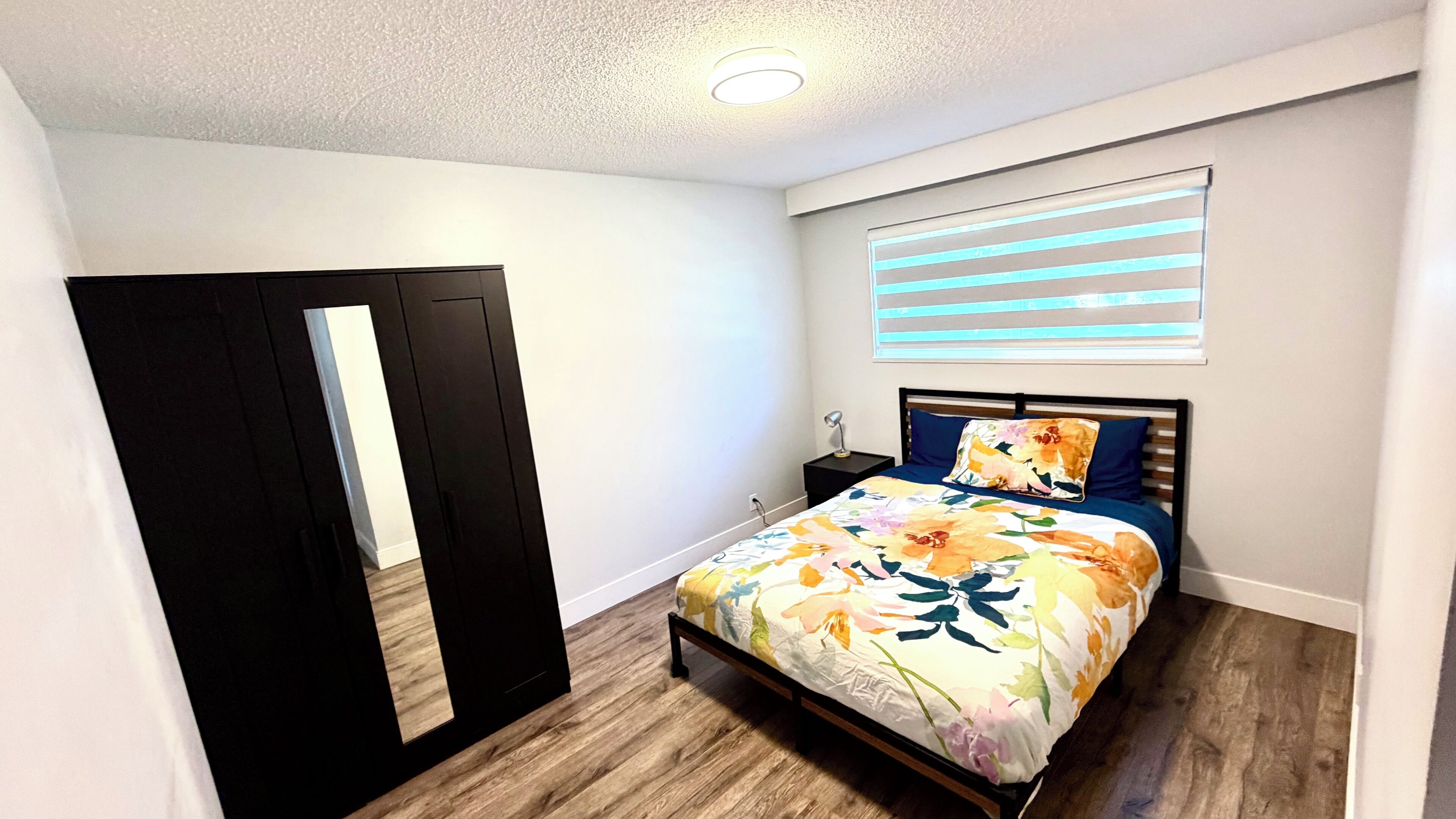 Un dormitorio con cama, mesita de noche, lámpara, ventana con persianas y puerta.