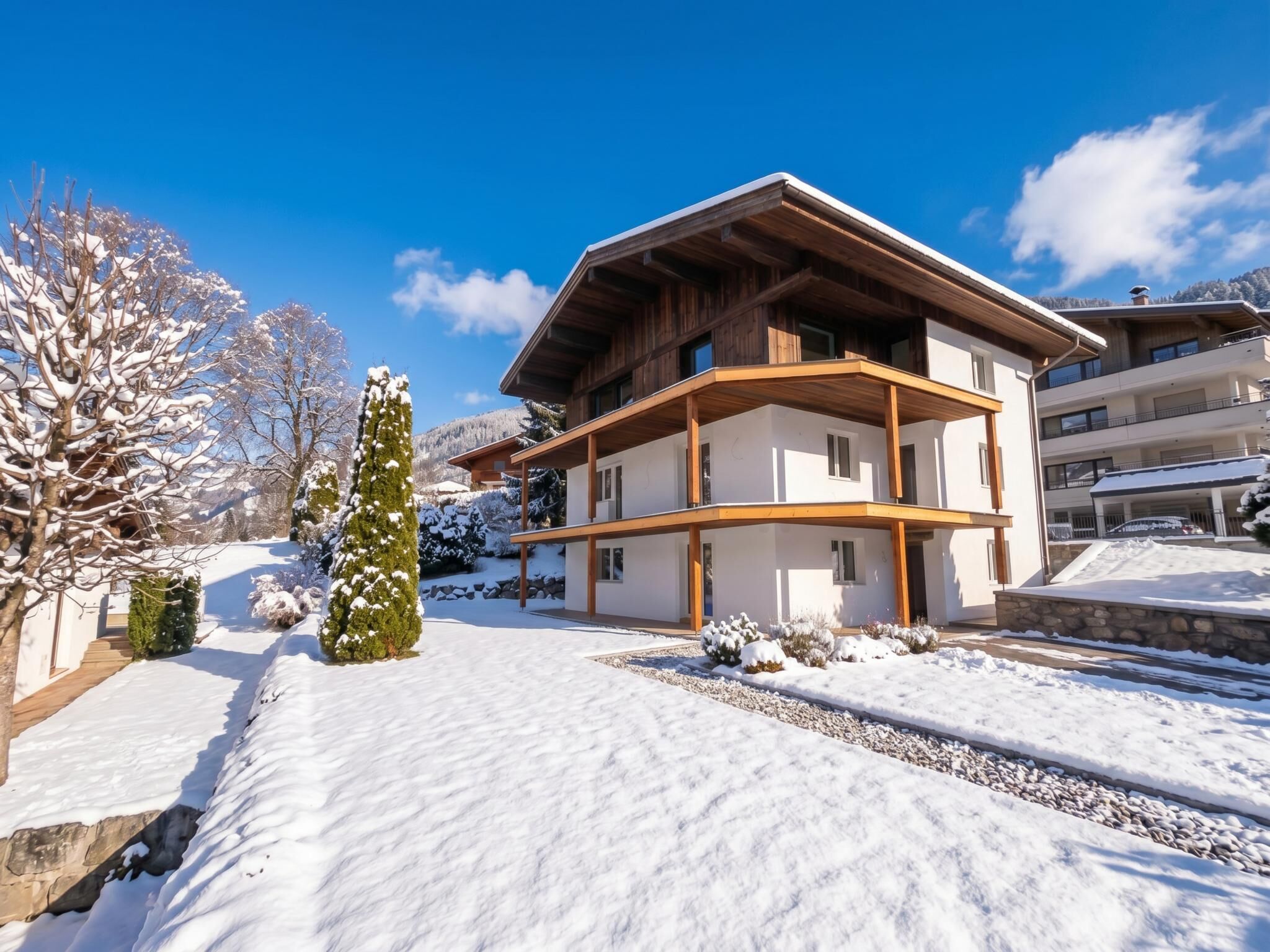 A modern two-story chalet with a wooden facade, a balcony, and a snow-covered landscape.
