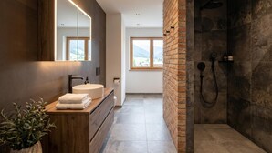 A modern bathroom with a wooden vanity, a large mirror, a round sink, and a shower area with a dark tile wall. - Stylish apartment with sauna, by the slopes (Brixen im Thale)