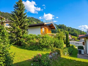 A modern house with a wooden deck, surrounded by lush greenery and a clear blue sky.