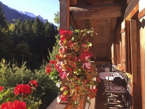 Ein Holzbalkon mit Tisch und Stühlen, umgeben von roten Blüten und Grünpflanzen, mit Blick auf eine gebirgige Landschaft.