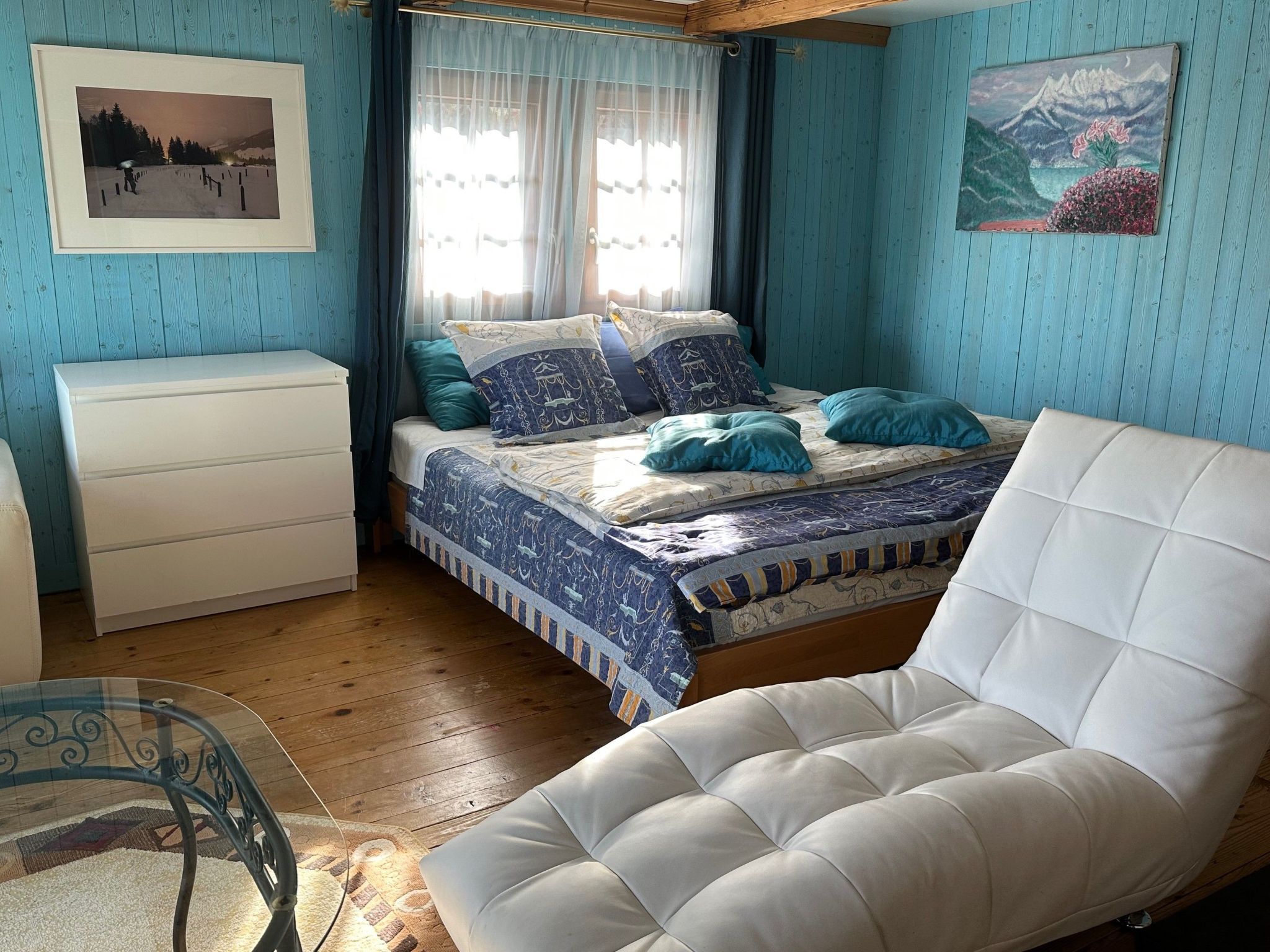 Une chambre à coucher avec un lit, une chaise blanche, une commode et une fenêtre avec des rideaux.