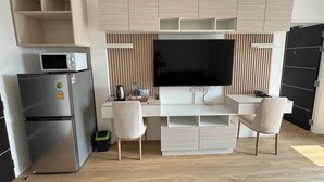 Una kitchenette moderna con microondas, refrigerador y televisor montado sobre un escritorio de madera con dos sillas.