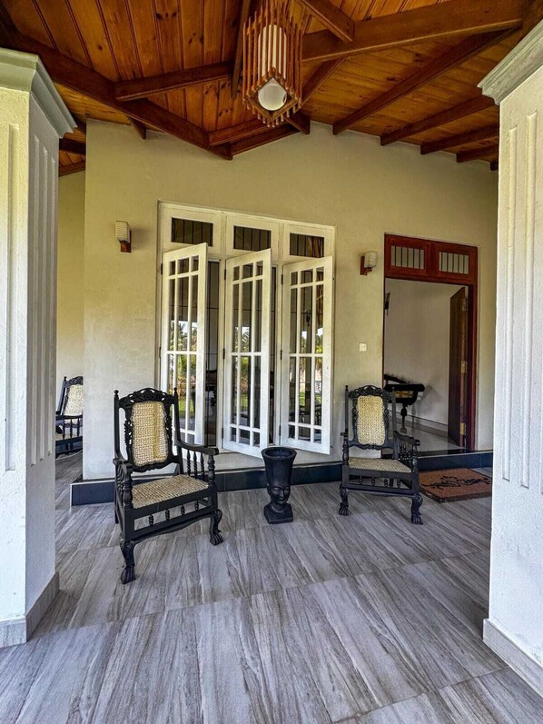 A covered porch with a wooden ceiling, two black chairs with woven seats, and a large white door with glass panels.