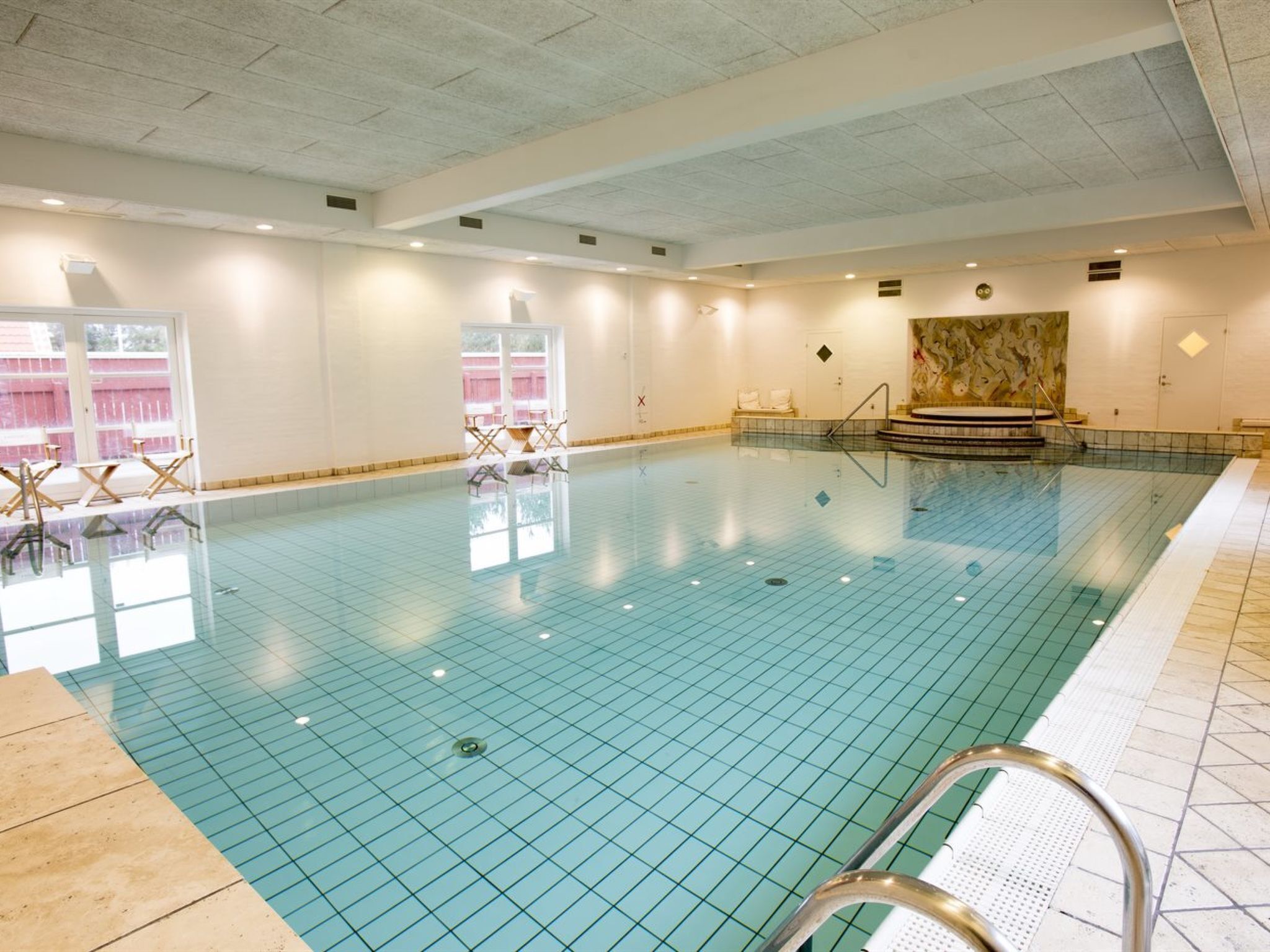 A large indoor swimming pool with tiled flooring, a diving board, and a seating area with chairs.