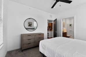 Ein Schlafzimmer mit einem Bett, einer Kommode, einem runden Spiegel, einem Deckenventilator und einer Tür zu einem Badezimmer.