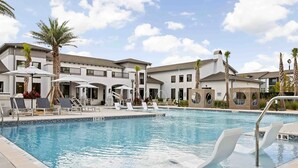 Una piscina con sillones de descanso y sombrillas, rodeada de edificios con balcones y palmeras.