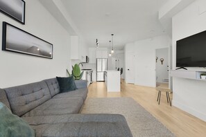 A modern living room with a grey sectional sofa, a flat-screen TV, and a kitchen in the background. - Spacious 1BR + Office in Central Langley (Langley Township)