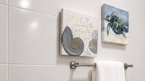 A bathroom with white tiles, a towel rack with a white towel, and two framed artworks on the wall.