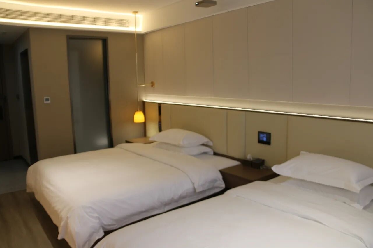 A hotel room with two beds, a bedside table, a lamp, and a wall-mounted control panel.