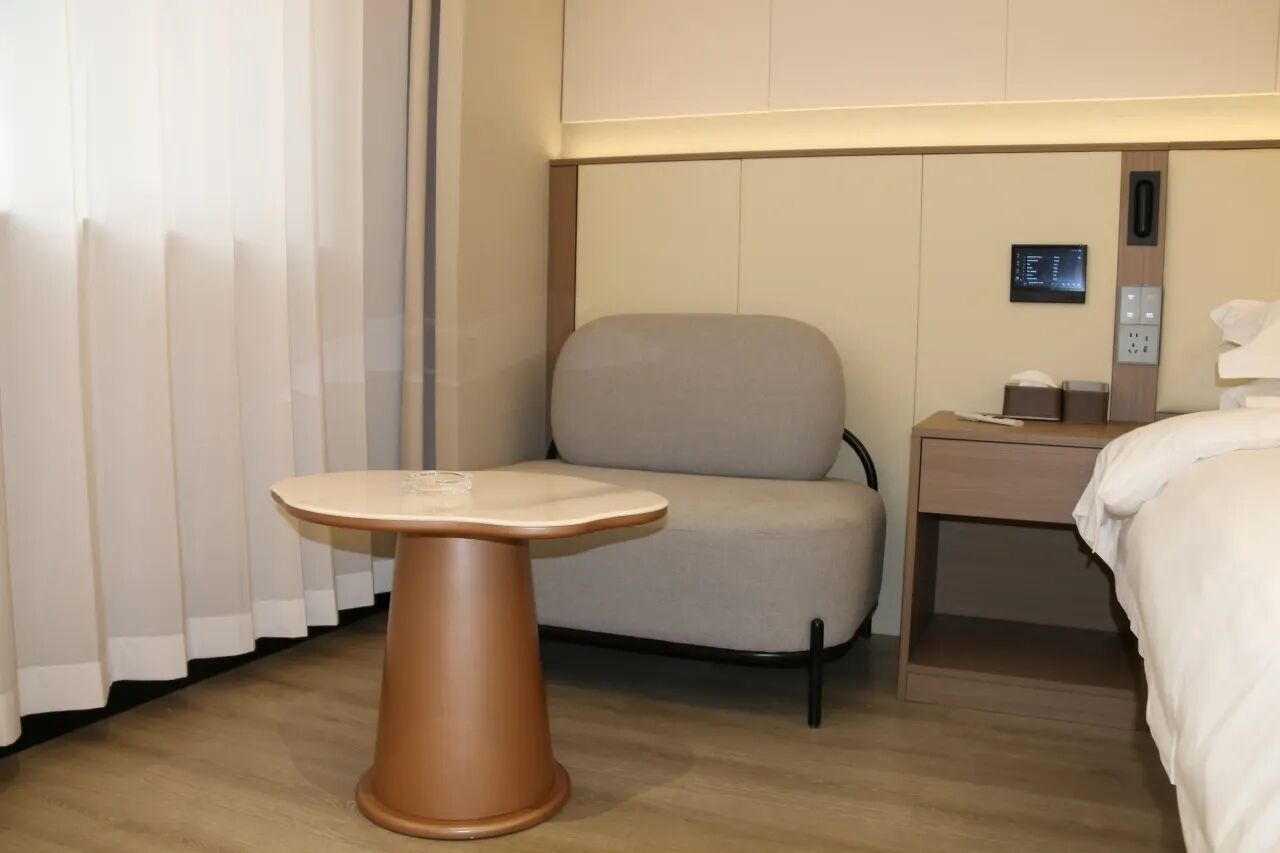 Habitación de hotel con cama, una silla, una mesita pequeña, una mesita de noche y un panel de control en la pared.