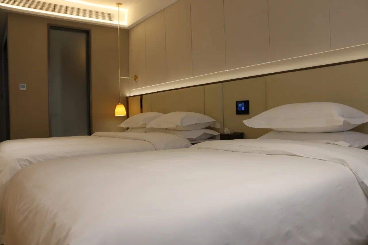Quarto de hotel com duas camas, uma mesinha de cabeceira, um abajur e um painel de controle fixado na parede.