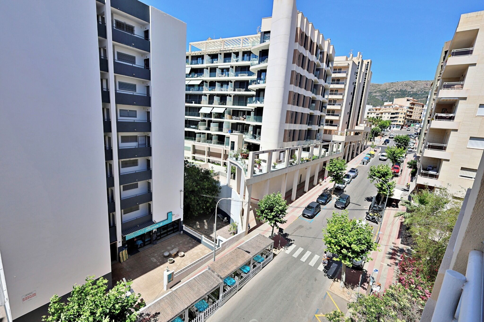 A multi-story residential building with balconies, a street view, and a clear sky.