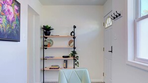 Una habitación con una mesa redonda, una estantería con objetos varios, una planta en maceta y una ventana.