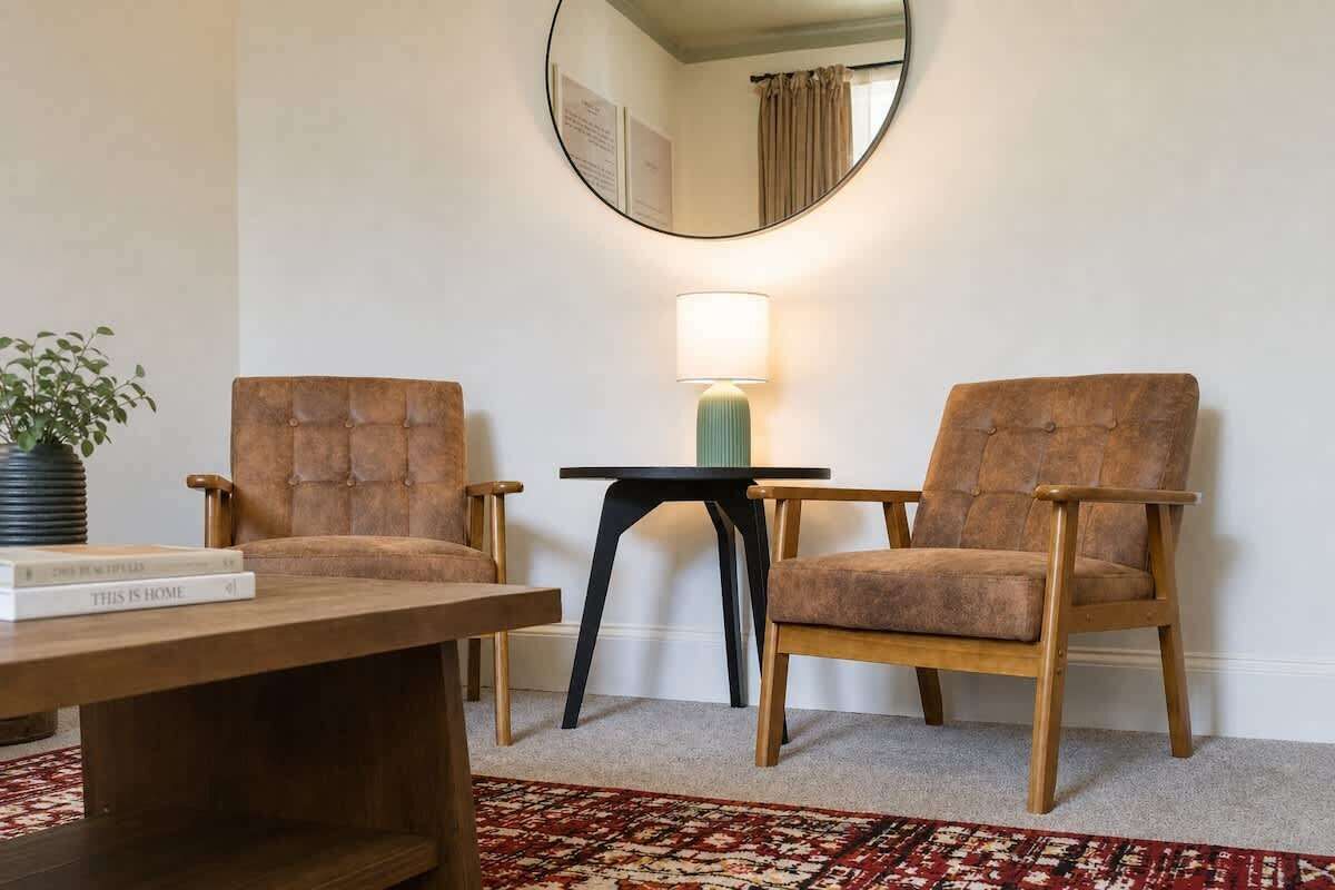 Um quarto com duas poltronas marrons estofadas, uma mesa pequena, uma luminária, um espelho e um tapete estampado.