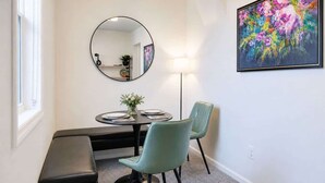 Un comedor con una mesa redonda, dos sillas, un banco, un espejo redondo, una lámpara de pie y un cuadro enmarcado en la pared.