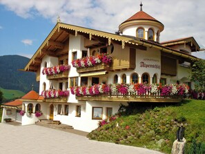 Ein mehrgeschossiges Gebäude mit Balkonen, die mit Blumen geschmückt sind, eine Schrifttafel mit der Aufschrift 'APPARTMENTS Alpenschlöfi' und ein bergiges Hintergrundpanorama.