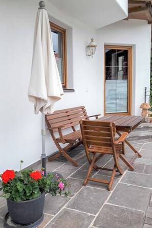 Ein Außenbereich mit Sitzgelegenheit, Tisch und Stühlen, einer Pflanze mit Blumen sowie einer Wandleuchte.
