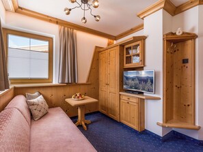 Ein gemütliches Zimmer mit einer Couch, einem kleinen Tisch mit Obst, Holzkästen, einem Fernseher und einem Fenster mit Vorhängen.
