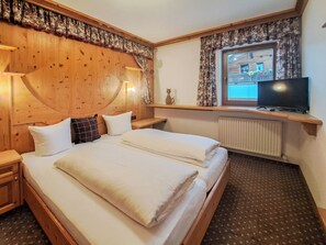 Een hotelkamer met een houten hoofdeinde, een bed met witte lakens, een nachtkastje, een televisie en een raam met gordijnen.