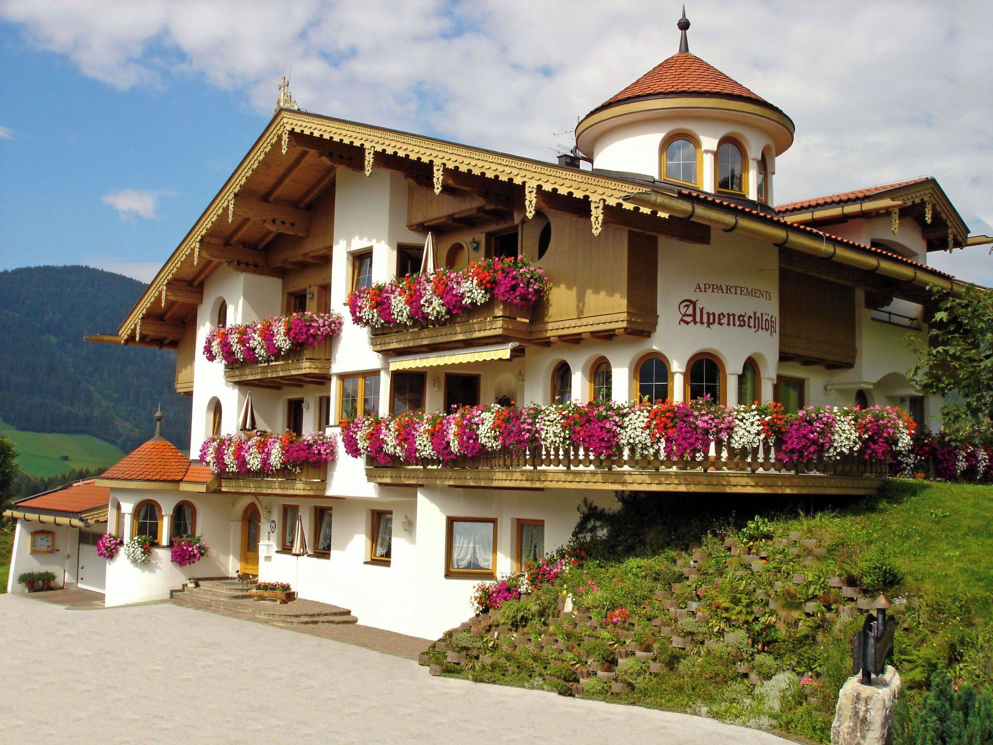 A multi-story building with balconies adorned with flowers, a sign reading 'APPARTMENTS Alpenschlöß', and a mountainous backdrop.