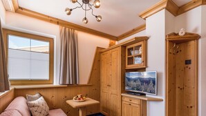 Ein gemütliches Zimmer mit einer Couch, einem kleinen Tisch mit Obst, Holzkästen, einem Fernseher und einem Fenster mit Vorhängen.