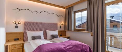 Ein Schlafzimmer mit einem hölzernen Kopfteil, einem großen Bett mit violetter Decke, zwei Nachttischen, einer Wandleuchte und einem Fenster mit Blick auf einen Berg.