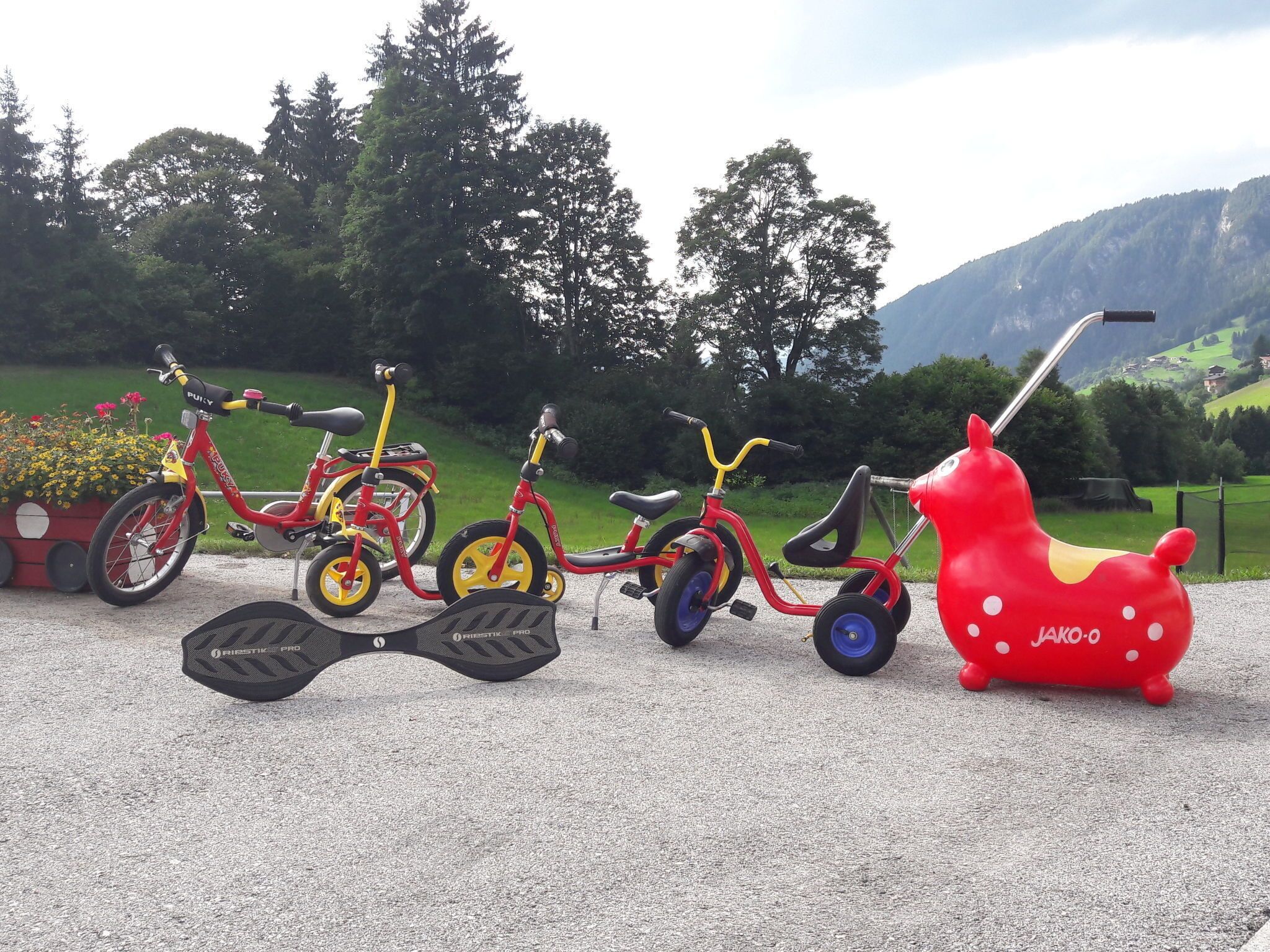 A row of children's tricycles and a red inflatable toy in a gravel area with greenery and mountains in the background.