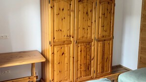 A wooden wardrobe with two doors, a wooden bench, and a bed with a light-colored blanket.