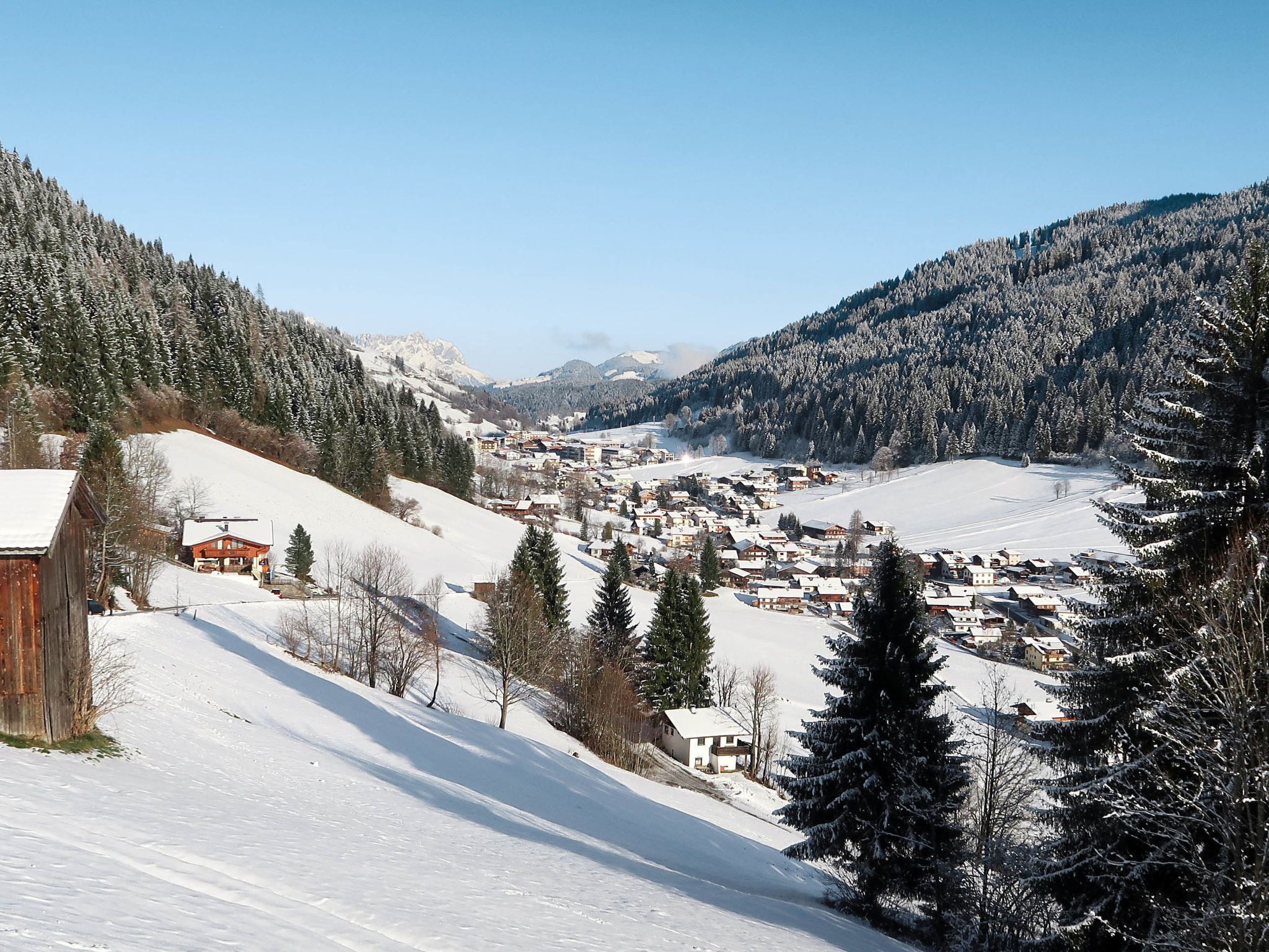 Ein schneebedecktes Dorf mit Häusern und einem Holzkonstrukt, umgeben von Bergen und Bäumen.