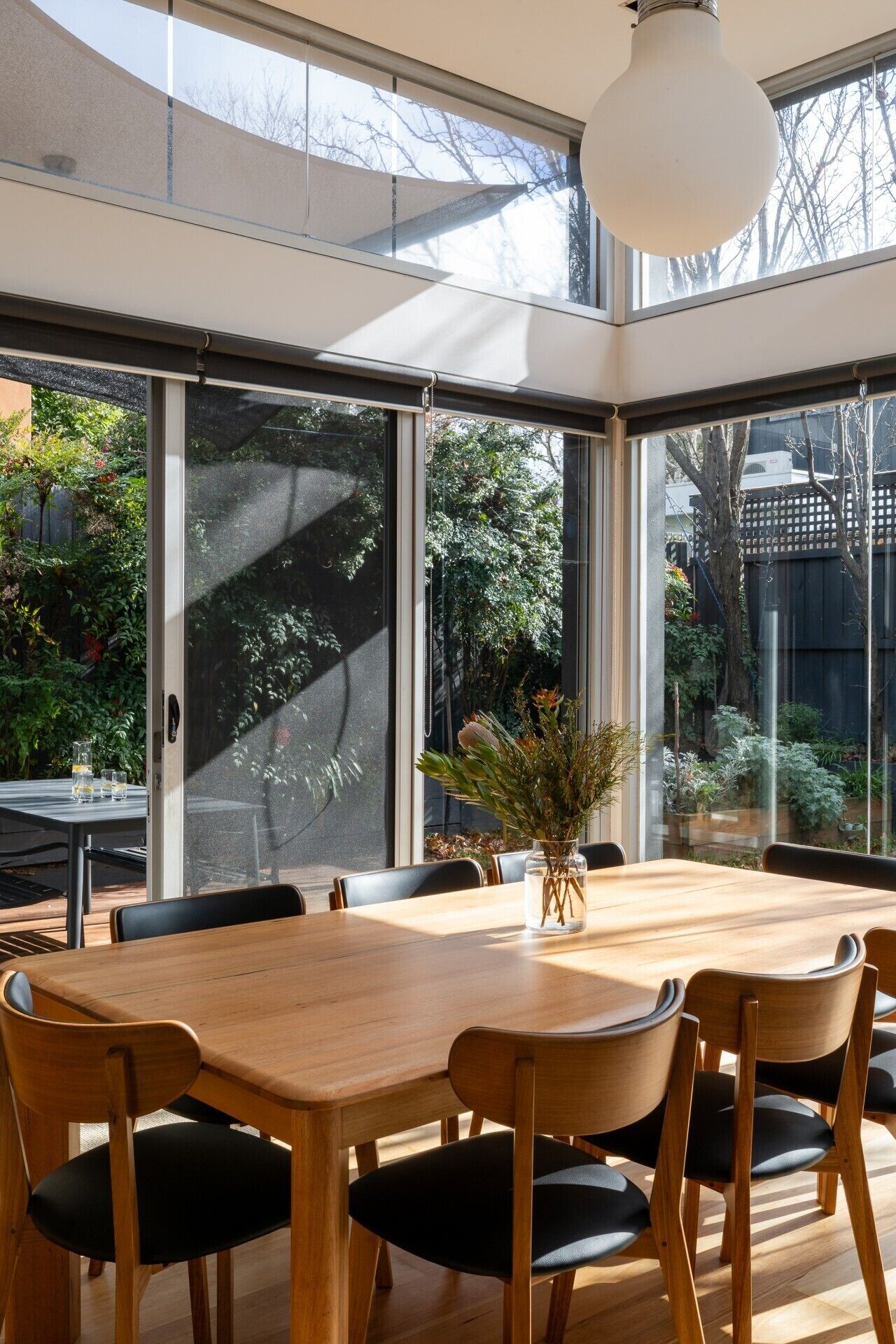 A modern dining area with a large wooden table, black cushioned chairs, and a vase with greenery.