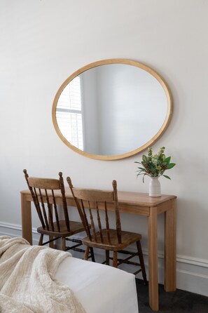 Een kamer met een houten bureau, een stoel, een ronde spiegel en een vaas met bloemen.