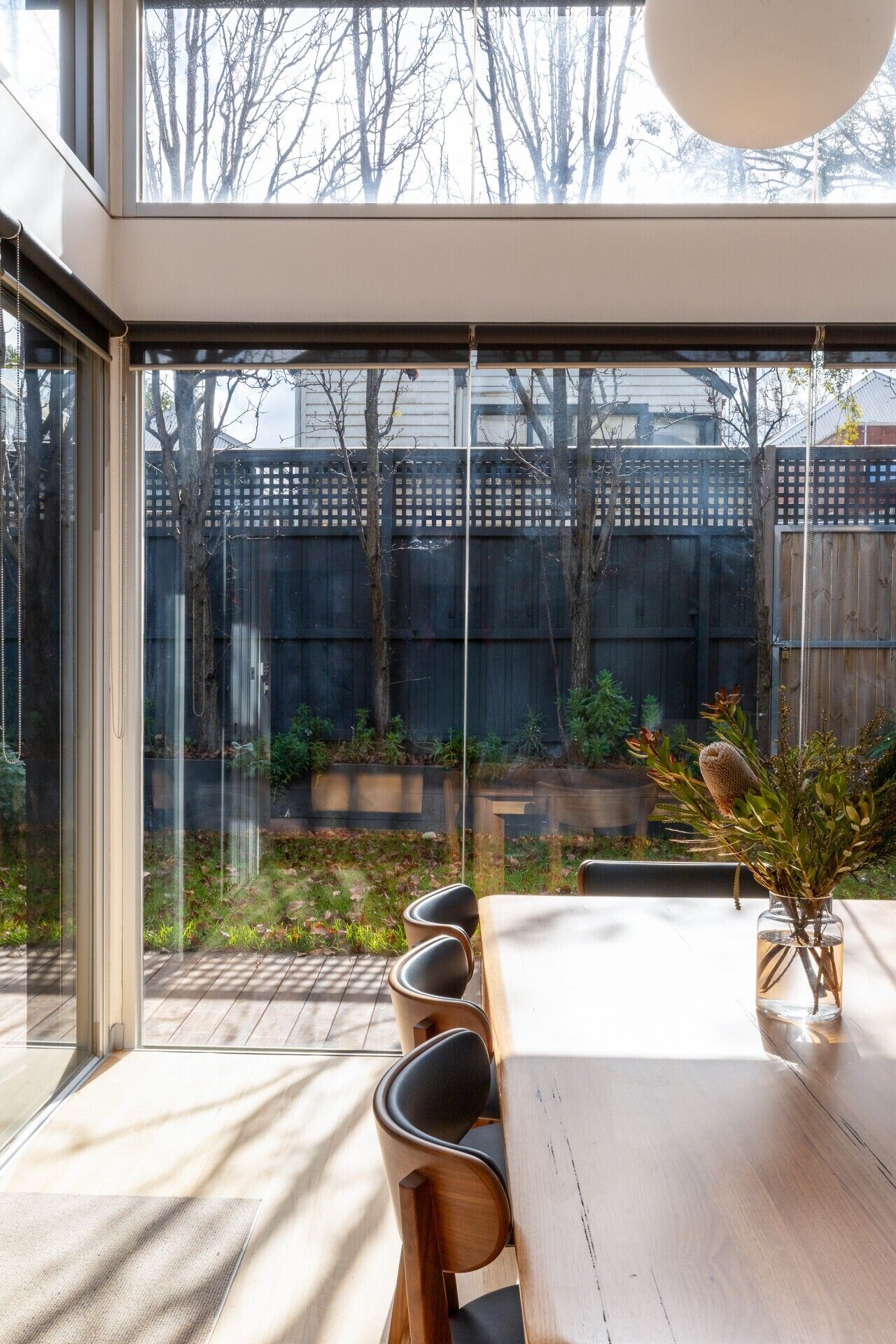 Een moderne eethoek met een grote tafel en stoelen, een potplant, en uitzicht op een achtertuin met een houten schutting en bomen.
