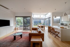 A modern living room with a sofa, a coffee table, and a TV. There is a dining area with a table and chairs. The room has large windows and a kitchen area with a microwave.